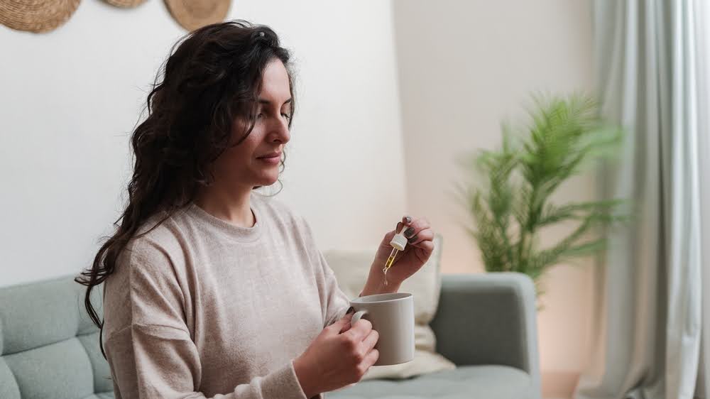 woman sitting on a couch dropping a solution from a tincture into a coffee mug