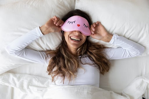 woman wearing all white and a pink sleep mask waking up rested with a smile on her face