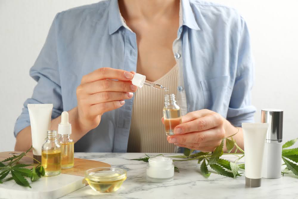 woman holding a tincture with a selection of creams and hemp leaves on the counter in front of her