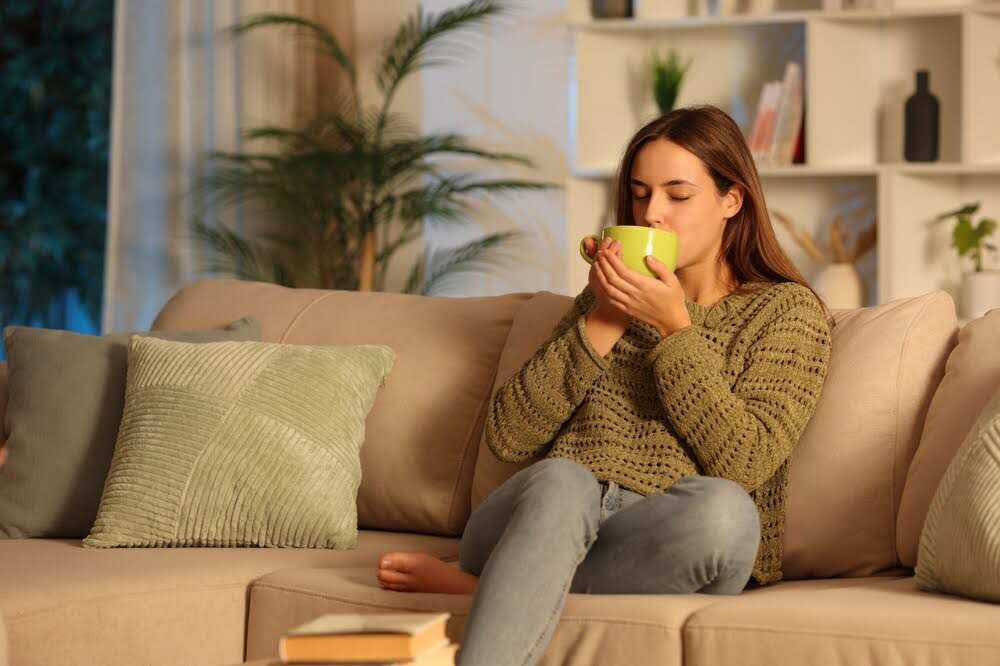 woman in a sweater drinking from a mug sitting on her couch