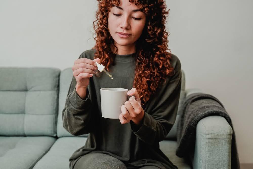 woman sitting on a couch and using a tincture dropper to add cbd oil to her drink mug