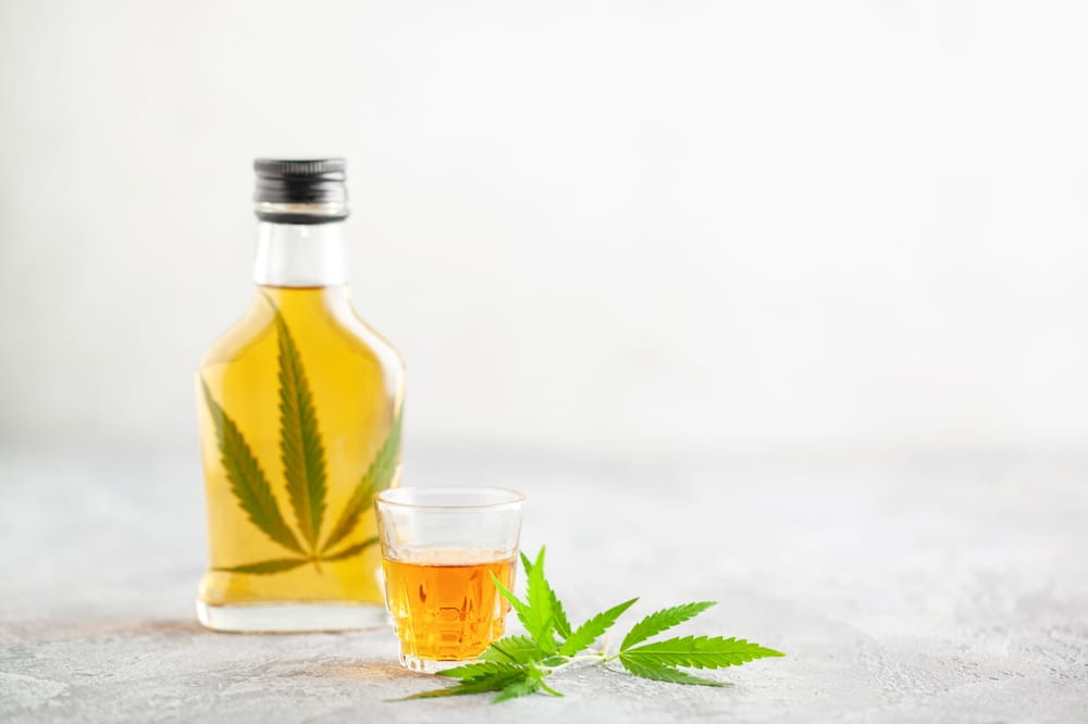 glass jar filled with an auburn liquid and a hemp leaf with a shot glass in front of it.