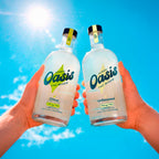 Two hands hold two clear glass bottles of Herbal Oasis The Mixer against a bright blue sky with sun rays. One bottle is labeled Citrus 10mg THC and the other is Unflavored 10mg THC.